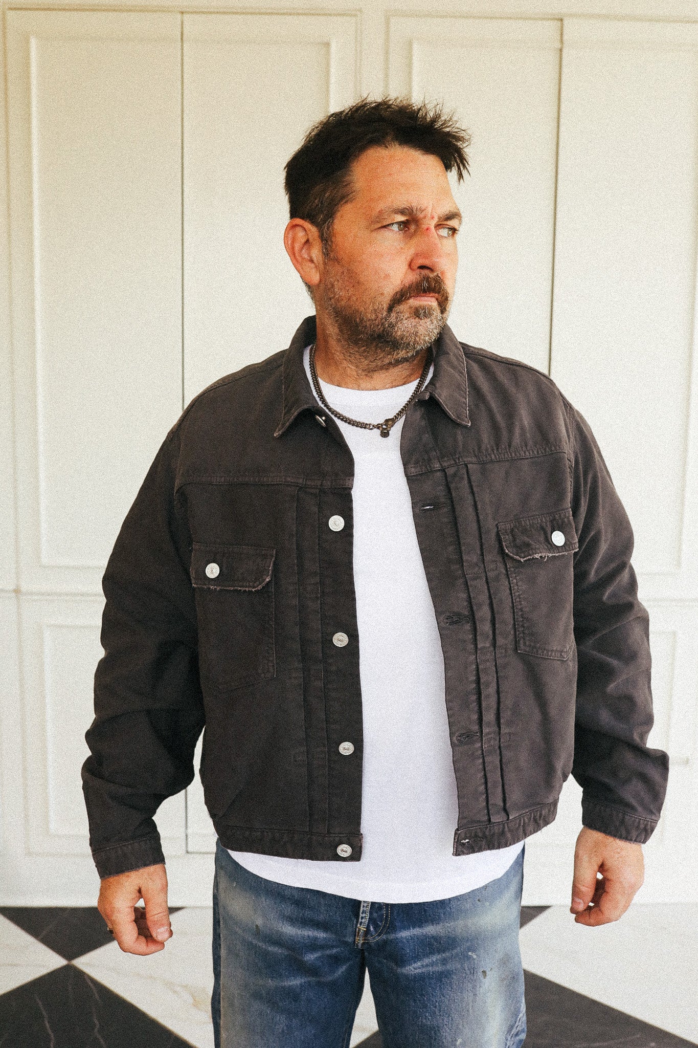 A man wearing a dark brown utility jacket over a white t-shirt and blue jeans, standing in front of a white paneled wall.