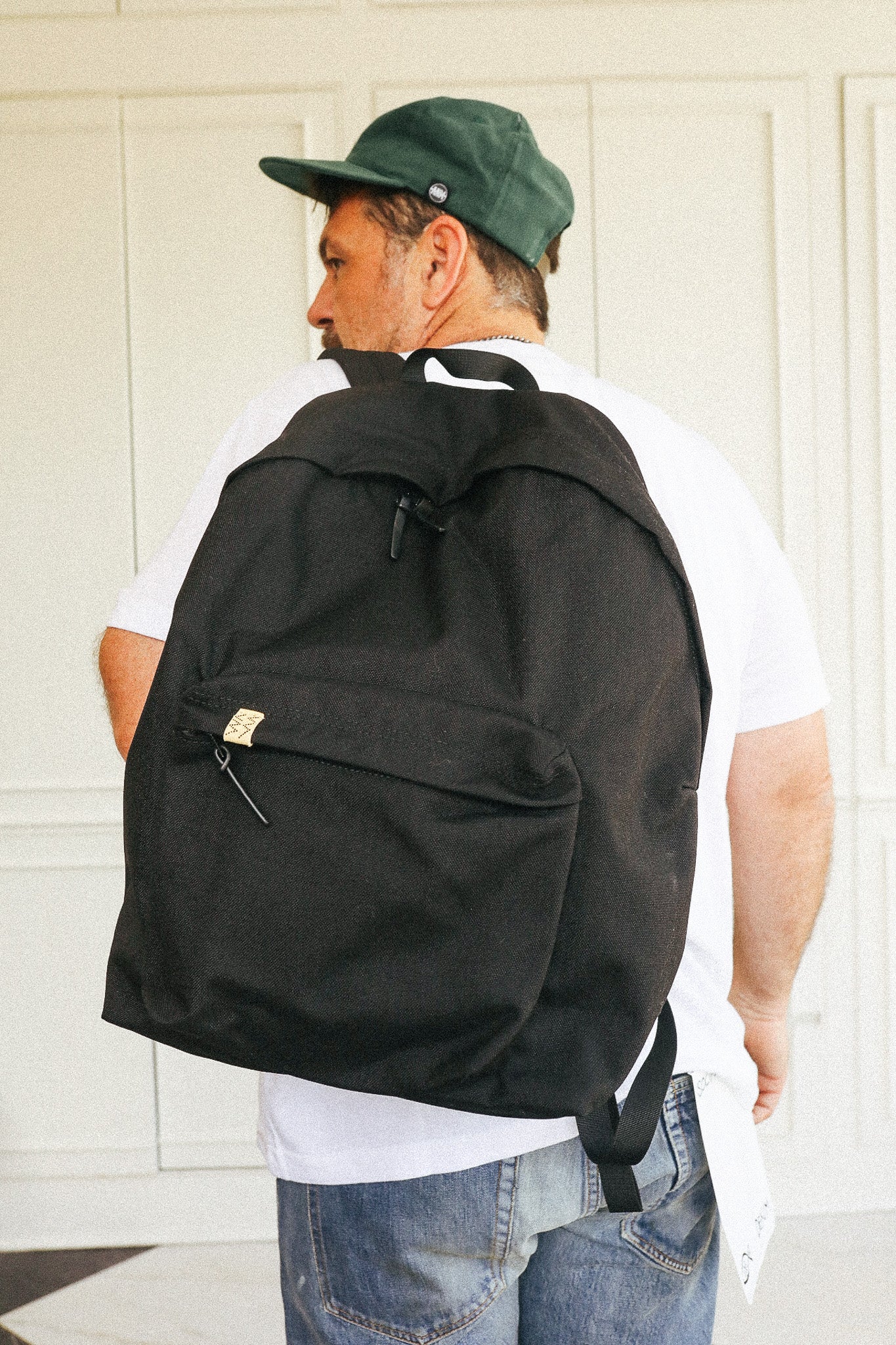 A man wearing a green cap, white t-shirt, and blue jeans is shown from the back, carrying a large black backpack with a front zippered pocket.