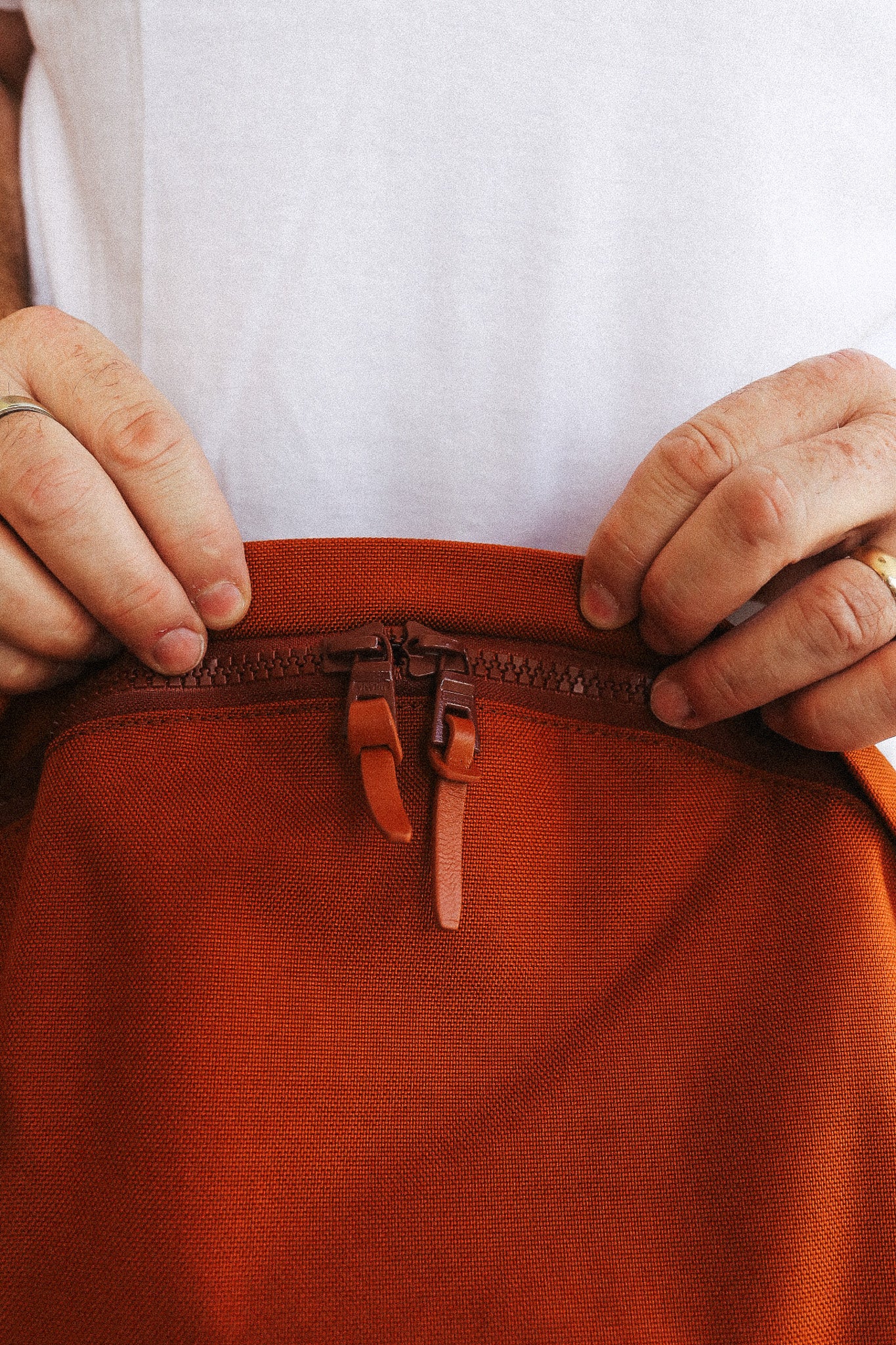 Close-up of a person's hands adjusting the zipper of a rust-orange canvas bag, wearing a white t-shirt.
