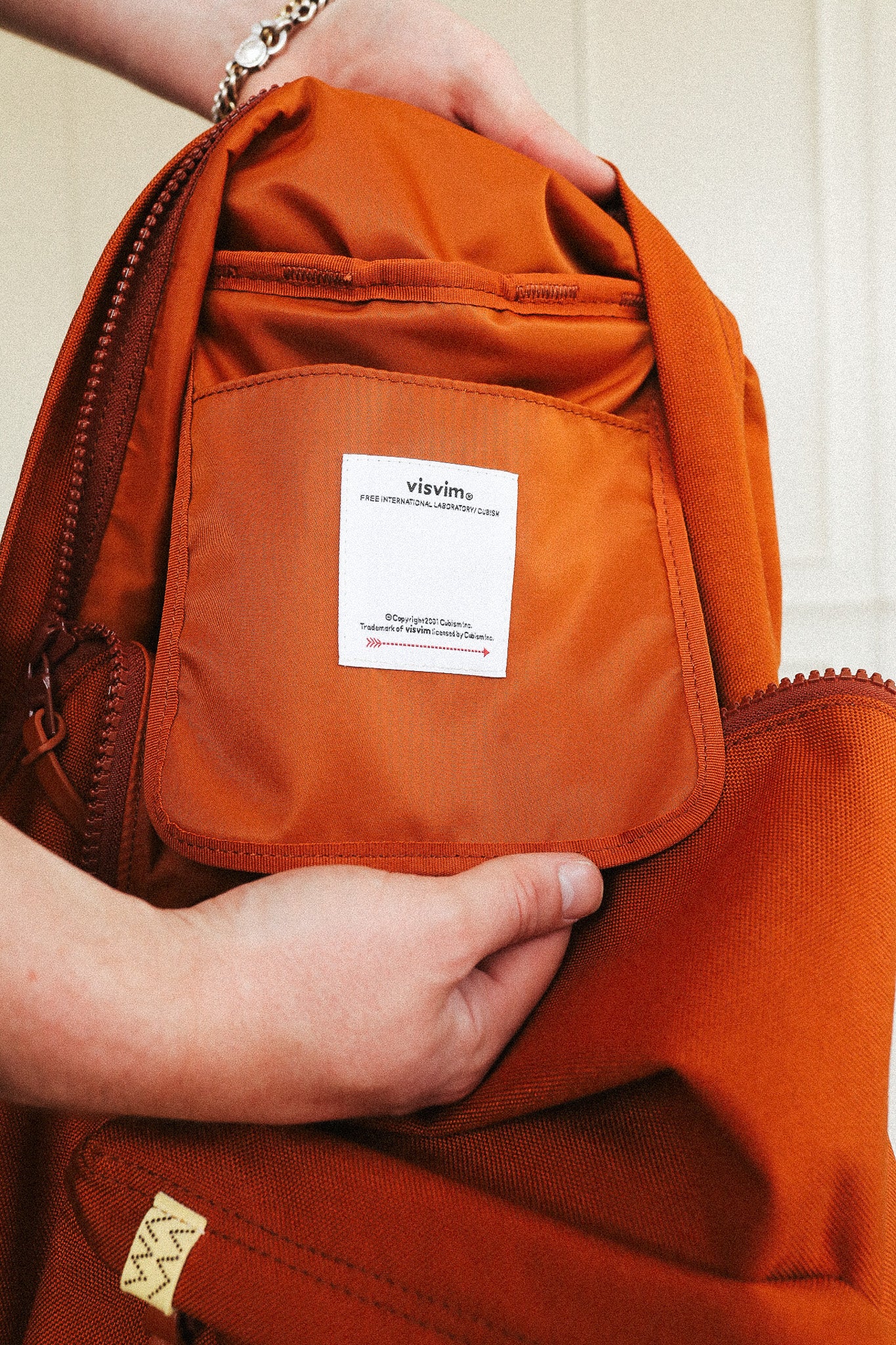 Close-up of a person holding an orange-brown nylon backpack, showing a white sewn-in label that reads 'viivima' on the front pocket.