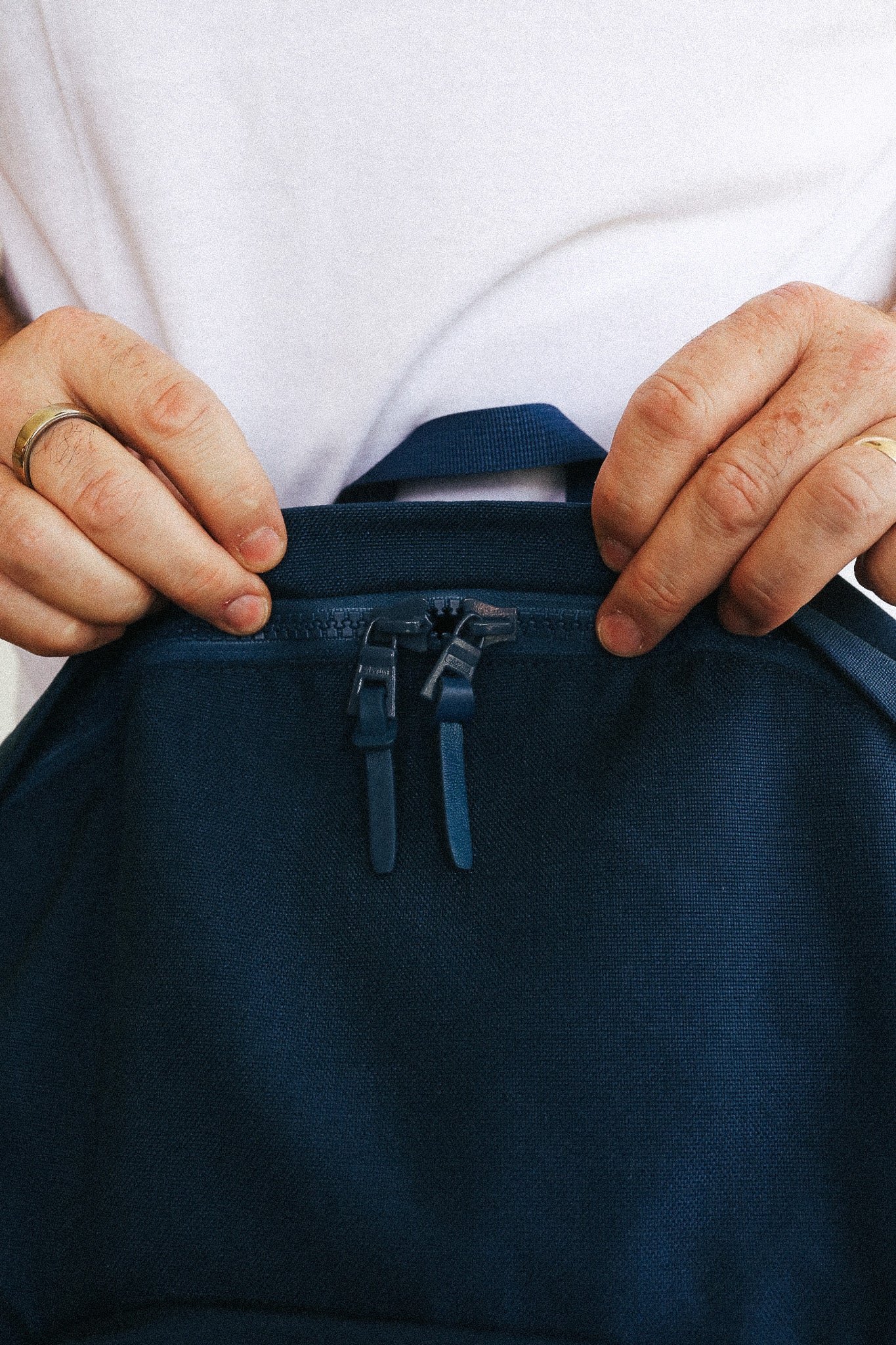 Close-up of a person's hands holding open a dark navy blue backpack or bag with a zipper pocket, wearing a white t-shirt.