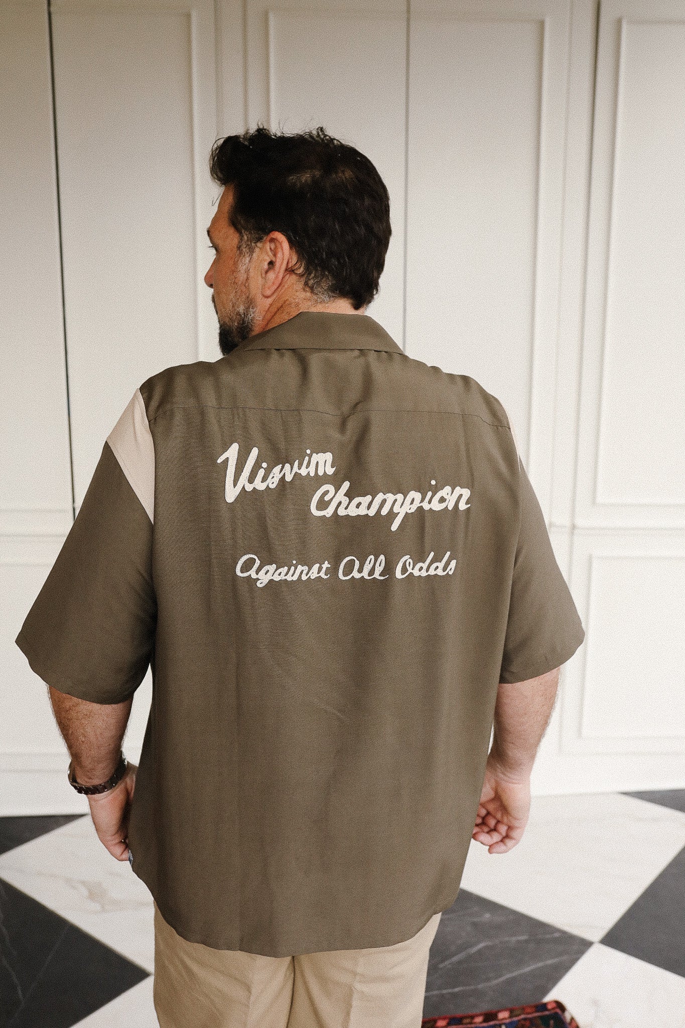 Man wearing a brown shirt with visible branding in a room with checkered floor.