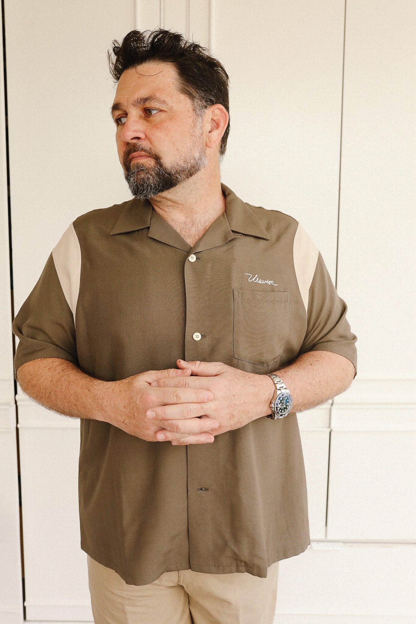 Man wearing a brown shirt with a logo, standing against a white wall.