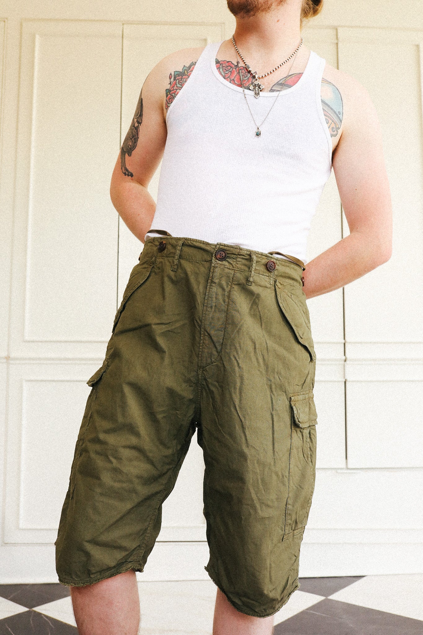 A person wearing olive green cargo shorts and a white tank top, standing in front of a white paneled door.