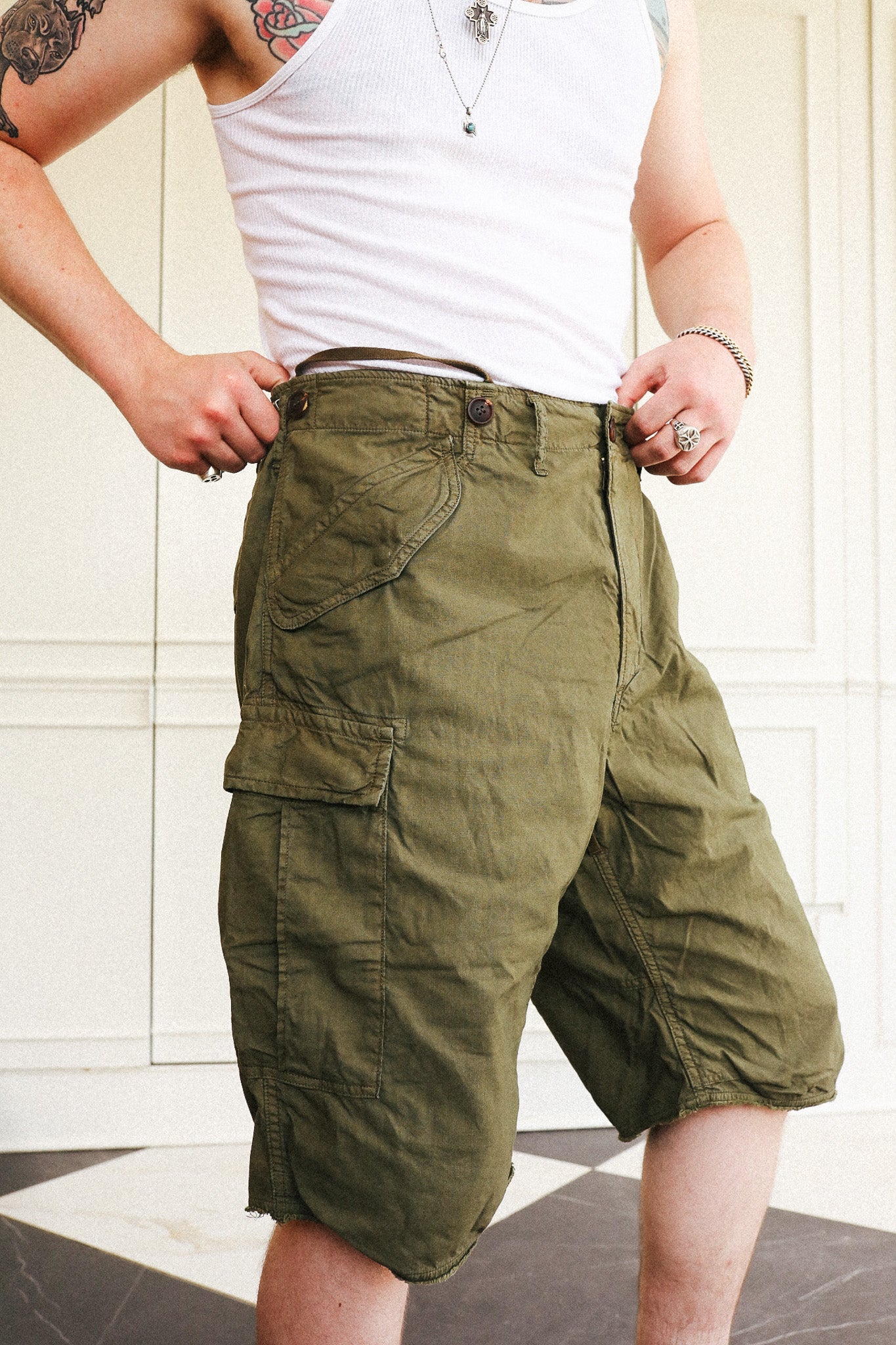 A person wearing olive green cargo shorts with a white ribbed tank top, standing in front of a white paneled wall.