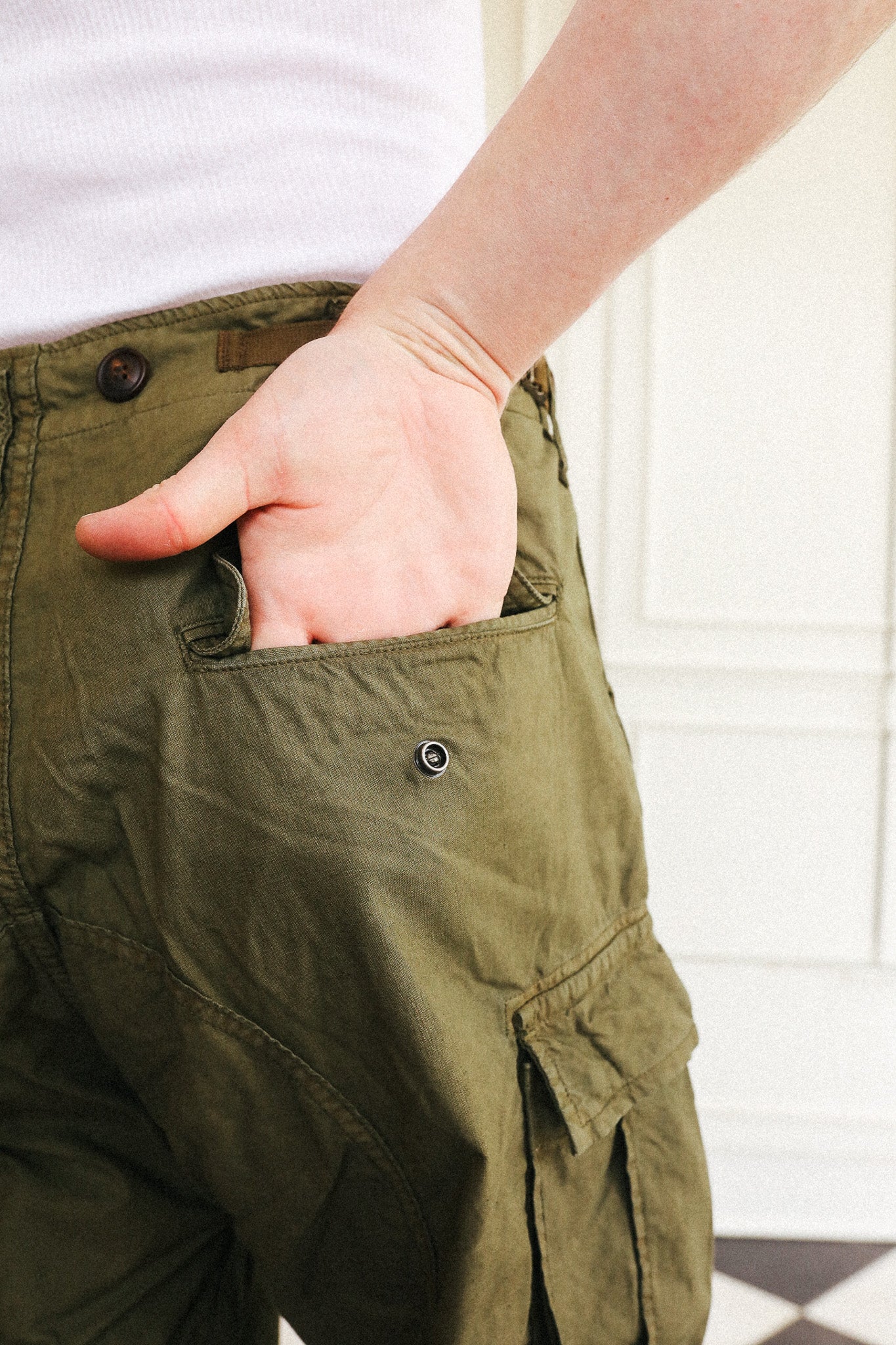 Close-up of a person wearing olive green cargo pants, with one hand tucked into the side pocket. The person is also wearing a white ribbed top, and the background shows a white paneled wall.