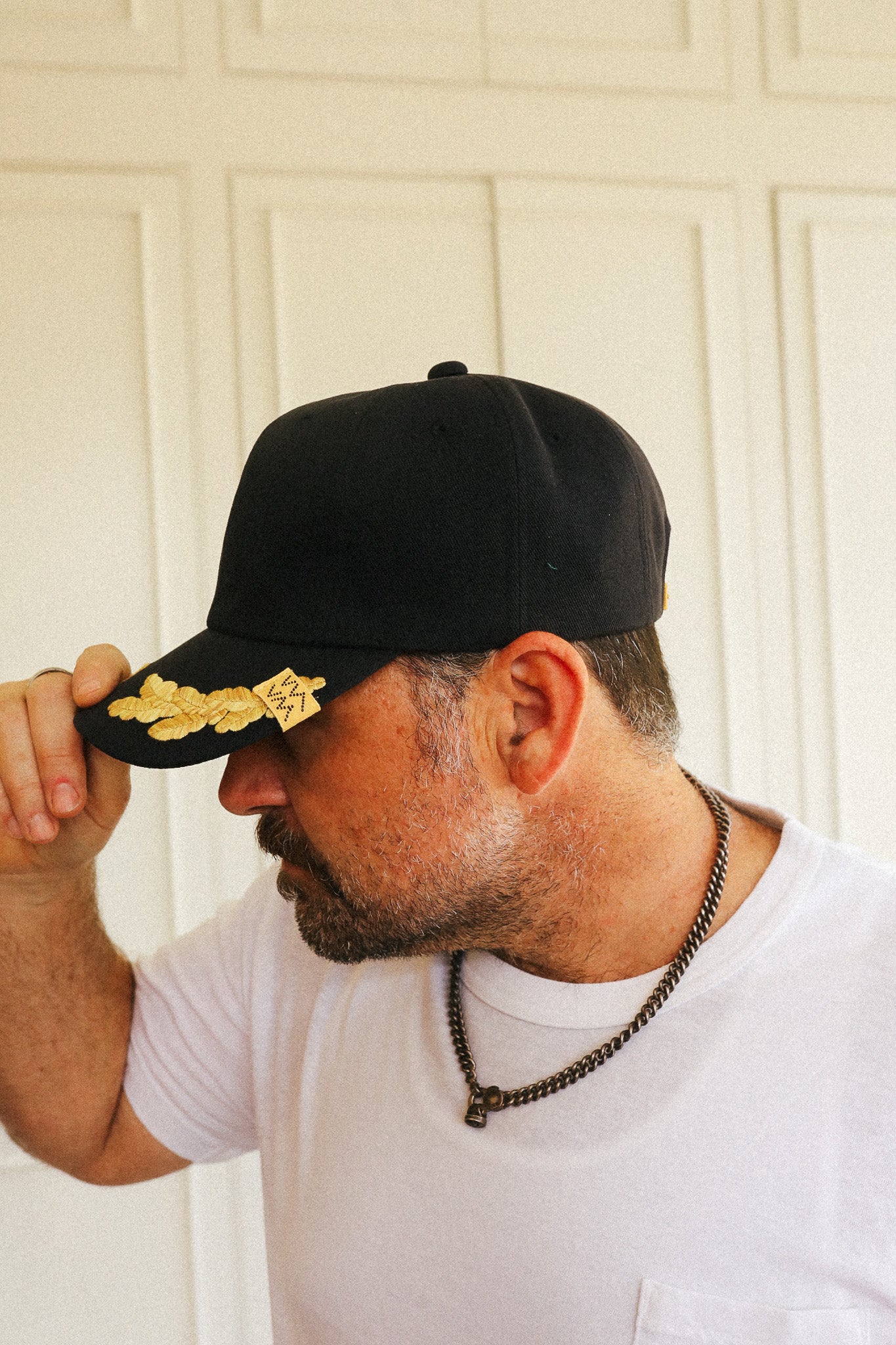 A man wearing a black baseball cap with a gold embroidered emblem on the brim, adjusting the brim with his hand. He is wearing a white t-shirt and a dark chain necklace, standing in front of a white paneled door.