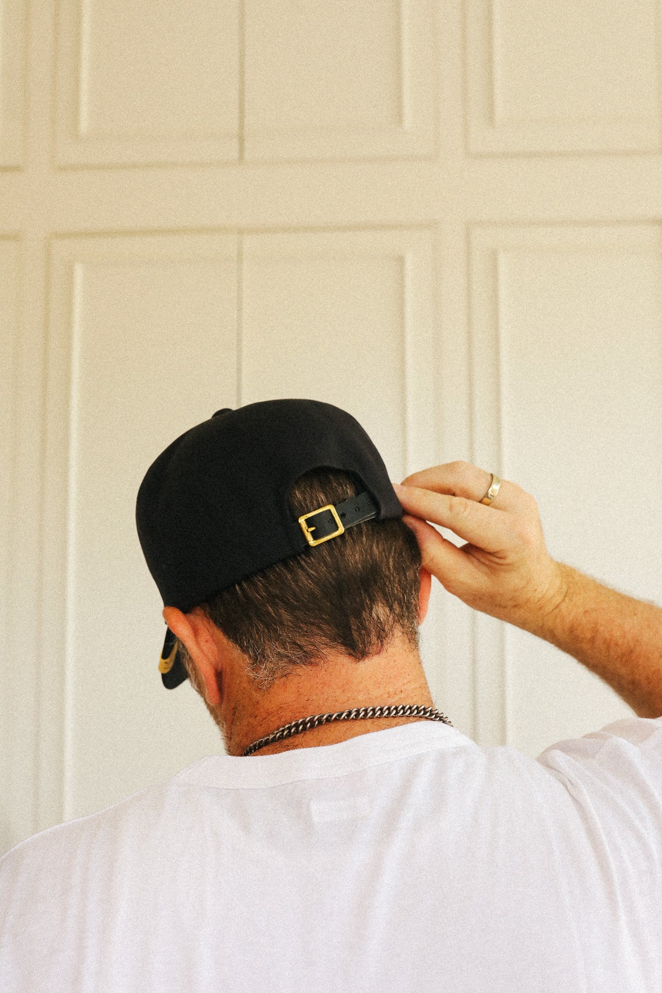 A man viewed from behind wearing a black baseball cap with a gold buckle, a white t-shirt, and a silver chain necklace, standing in front of a cream-colored paneled door.