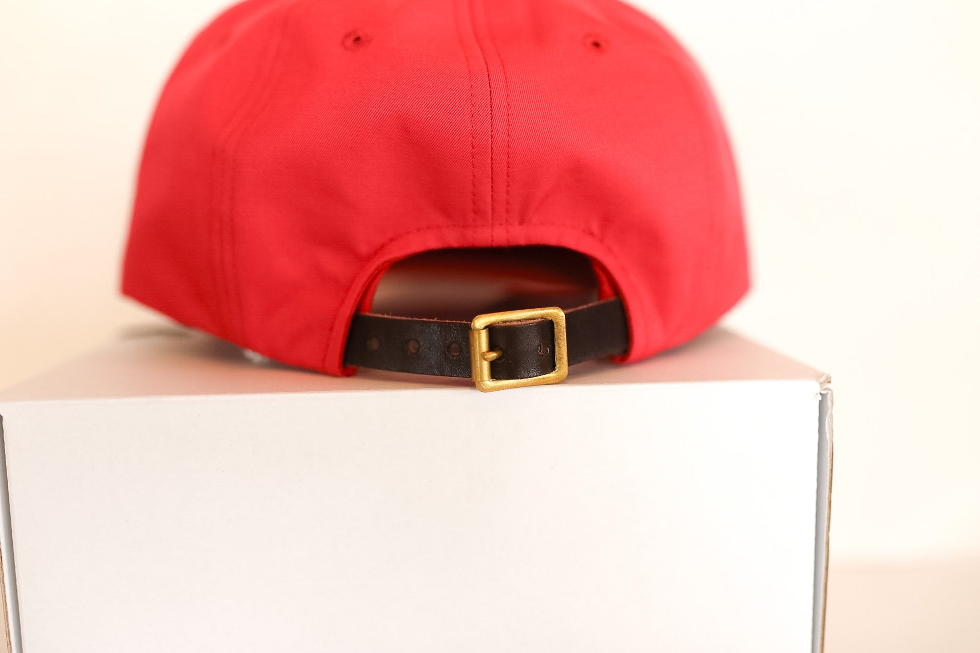 Red cap with a brown belt and gold buckle on a white box against a beige background