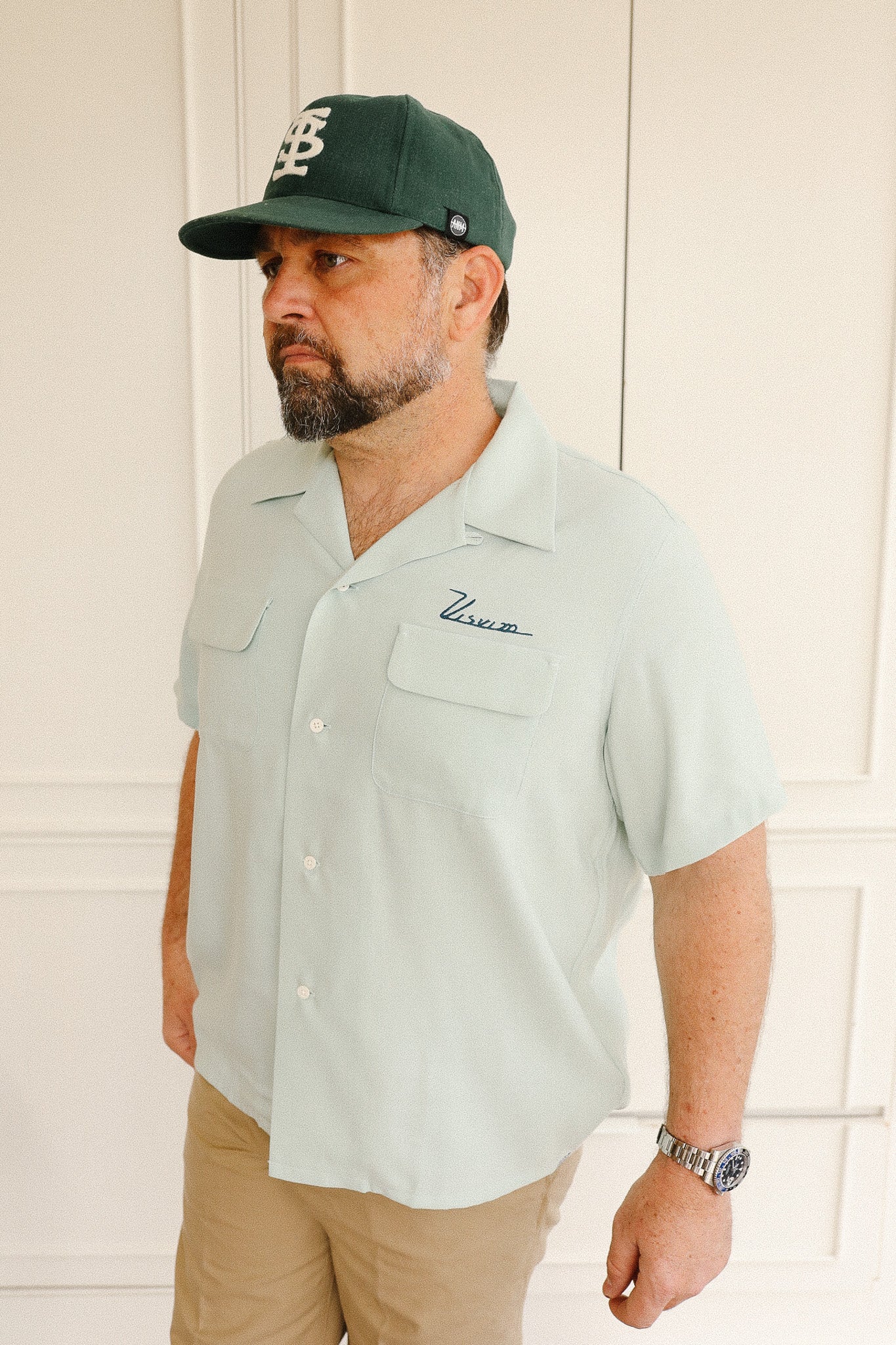 Man wearing a light green button-up shirt with a logo, beige pants, and a green cap indoors.