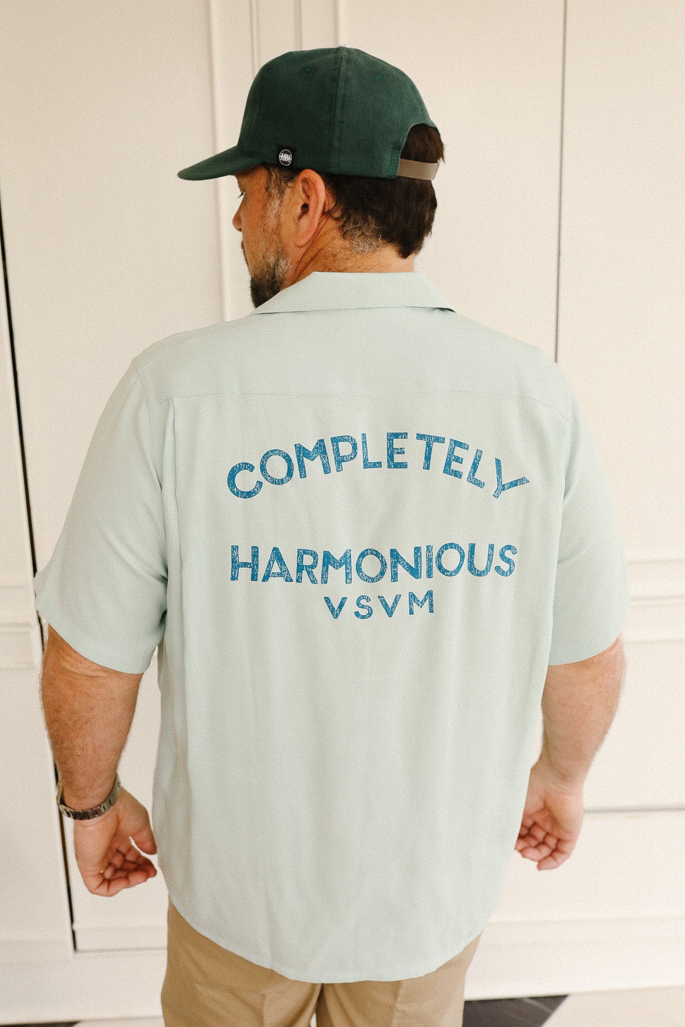 Man wearing a light green shirt with text on the back, standing against a white wall.