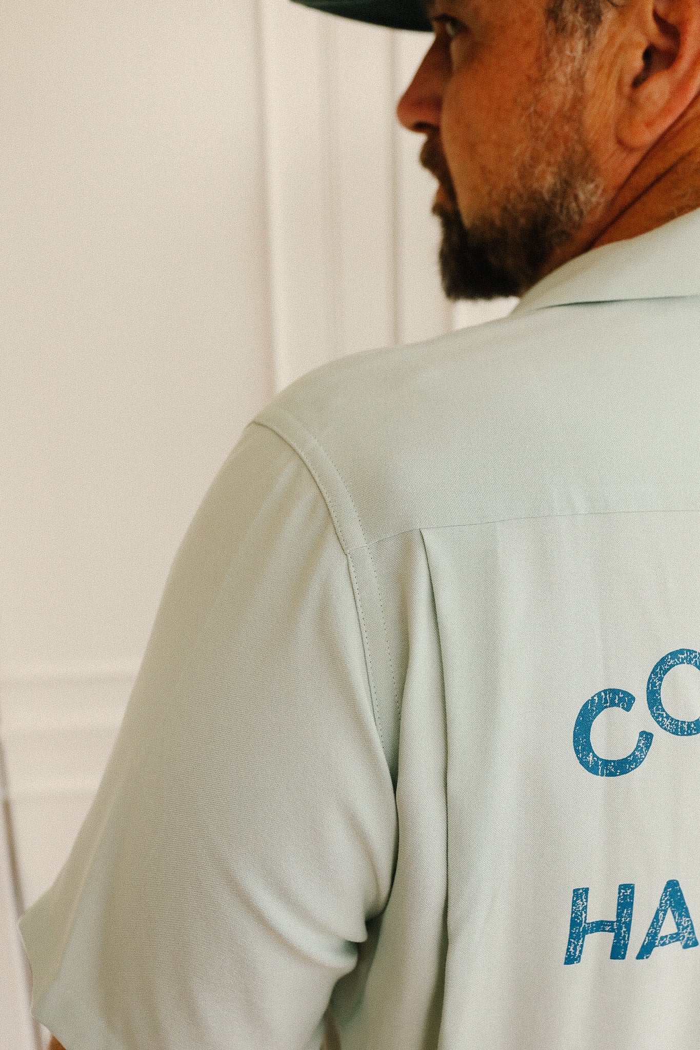 Man wearing a light green shirt with text on the back, standing against a white curtain.