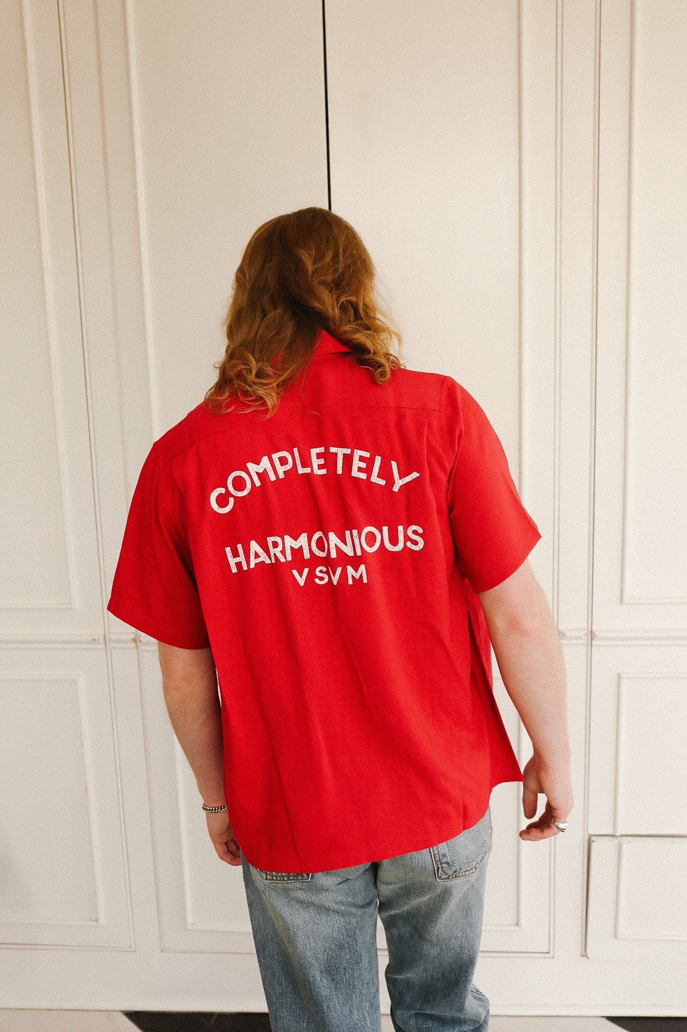 Person wearing a red shirt with text on the back, standing against a white paneled wall.