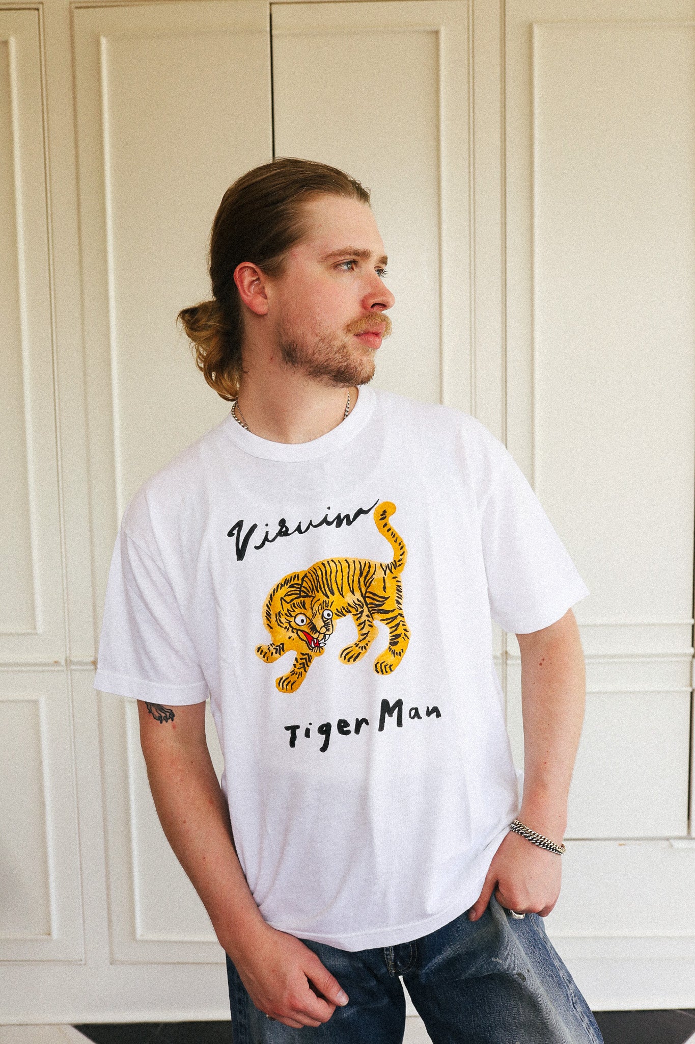 A man wearing a white t-shirt with a yellow and black tiger graphic and the text 'Vibissim Tiger Man' printed on the front, standing in front of a cream-colored paneled door.