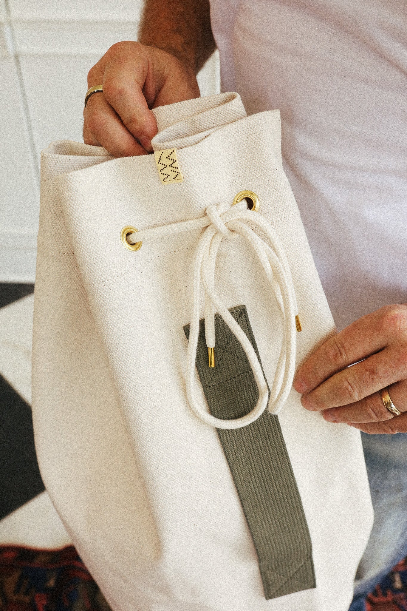 A close-up of a cream canvas drawstring backpack being held by a person. The bag features white drawstrings threaded through gold-tone metal grommets, a small gold geometric logo patch near the top, and a vertical olive green fabric accent panel on the front.