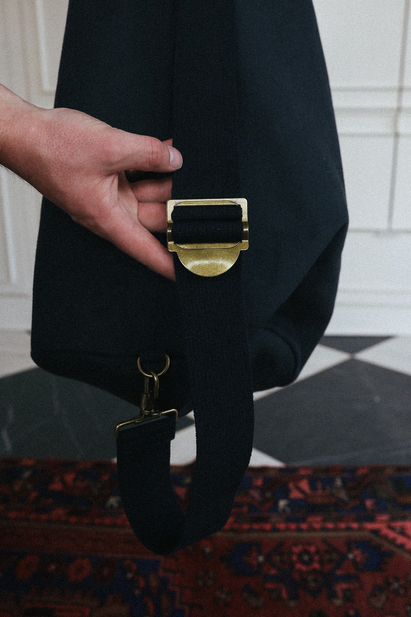 A close-up of a person's hand holding a black shoulder bag with a wide, adjustable black strap and a prominent gold-tone metal buckle, set against an indoor background with a patterned rug.