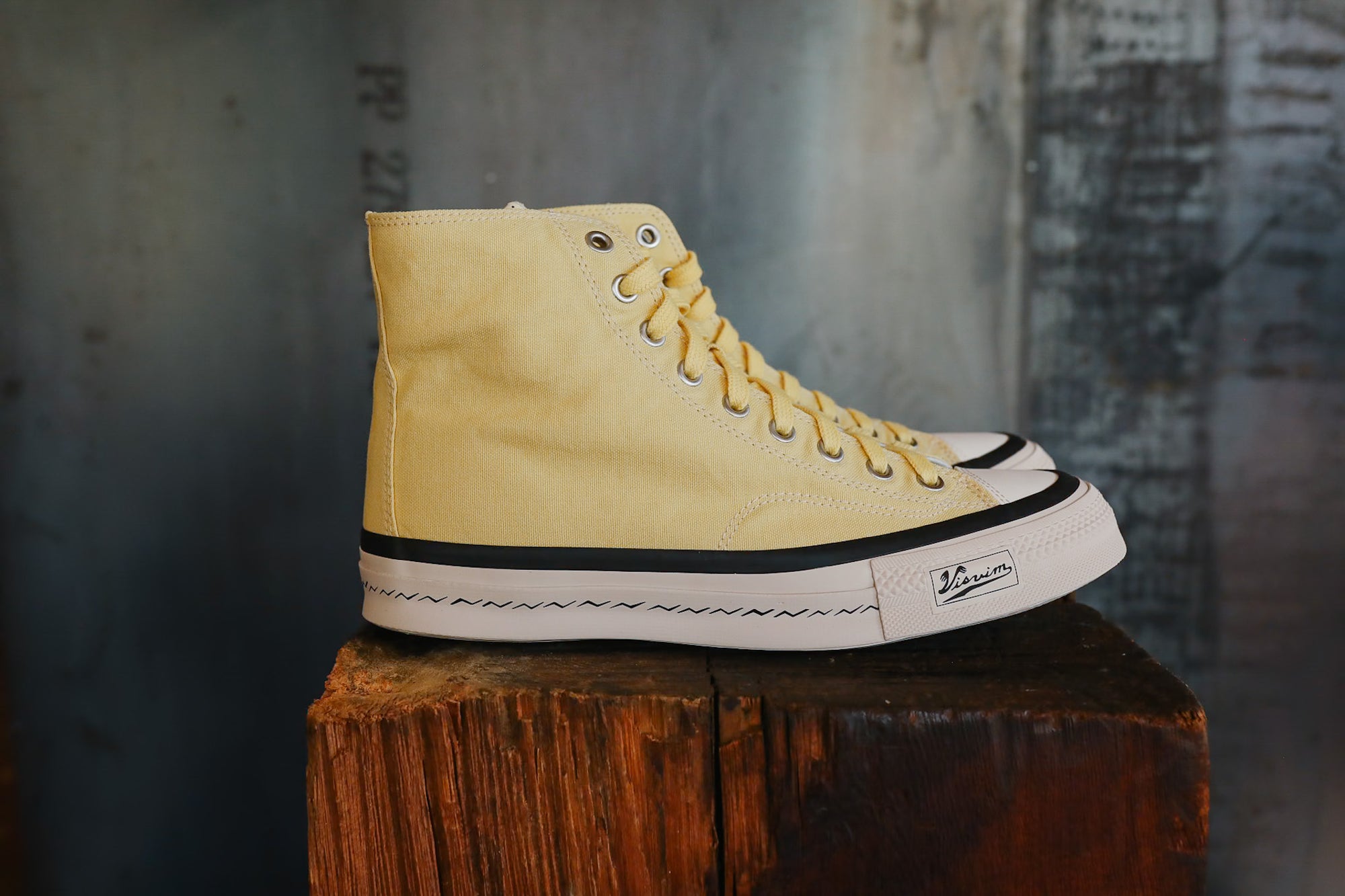 Yellow high-top sneaker with white laces on a wooden block against a textured wall.