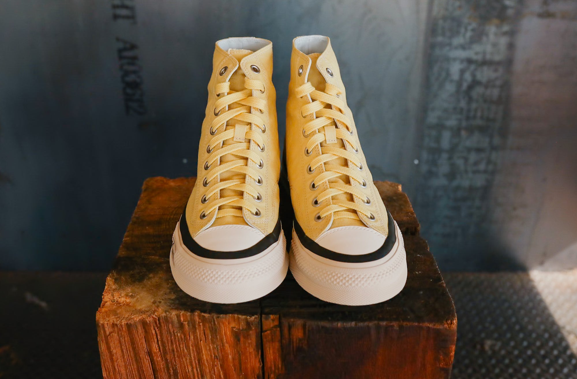 Pair of yellow high-top sneakers with black soles on a wooden block against a textured wall.