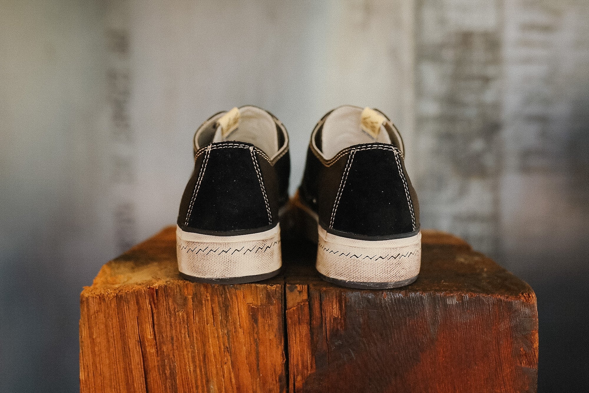 Pair of black sneakers with white soles on a wooden block against a textured wall.