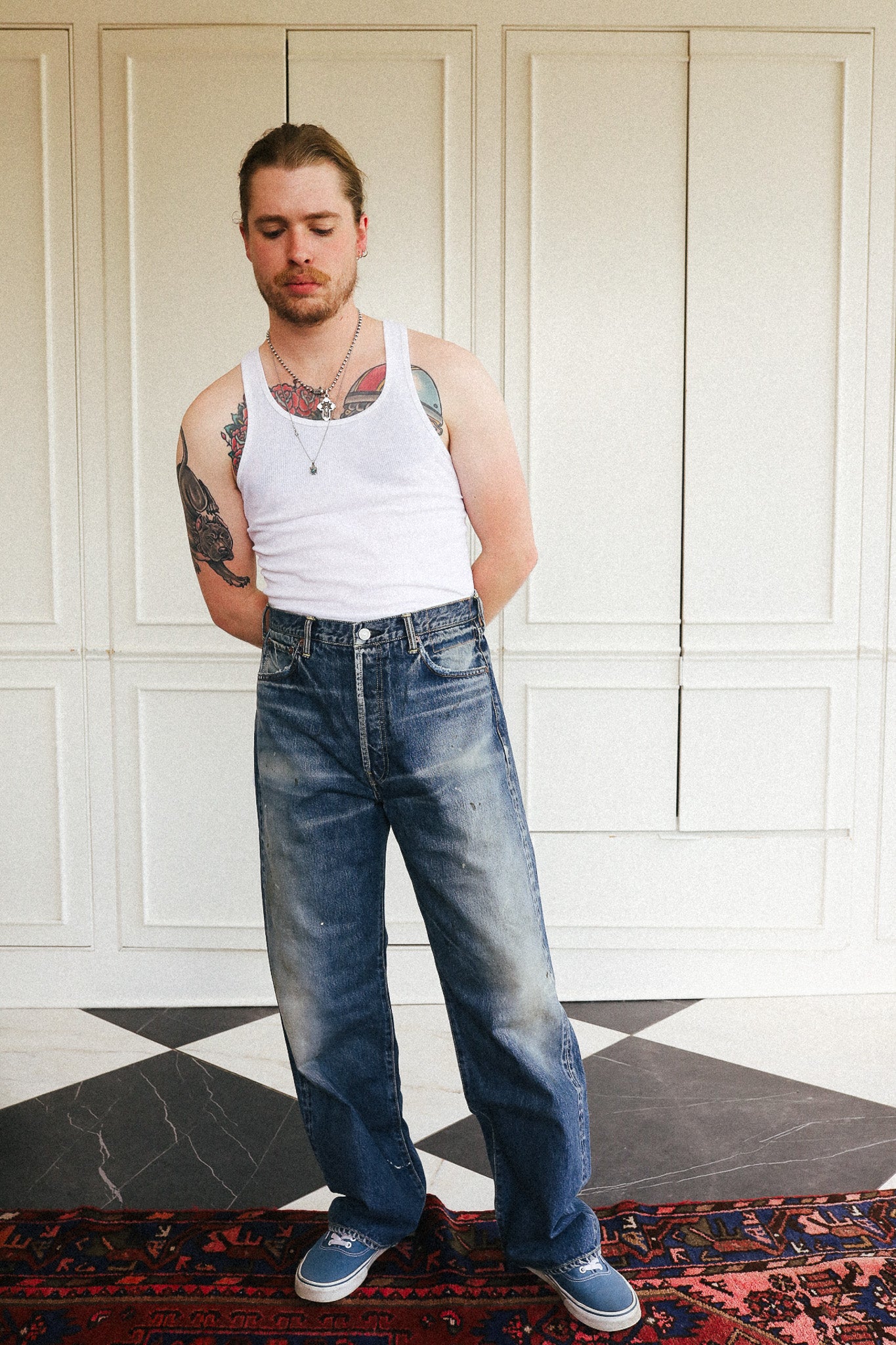 A man wearing a white tank top, wide-leg blue jeans, and blue sneakers, standing in front of white paneled doors.
