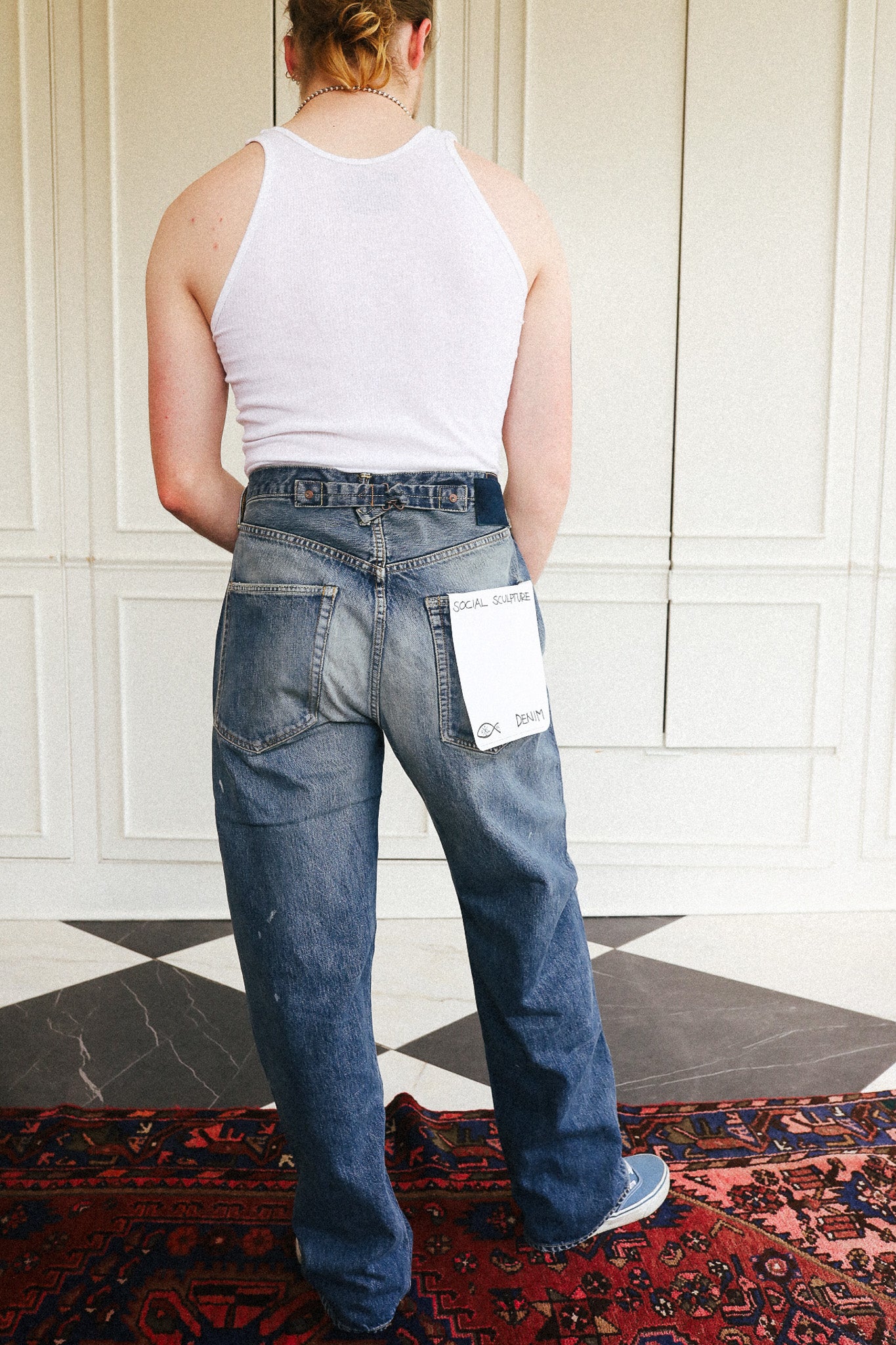 A person viewed from behind wearing blue wide-leg jeans with a white tank top, standing indoors against a white paneled wall on a patterned rug.