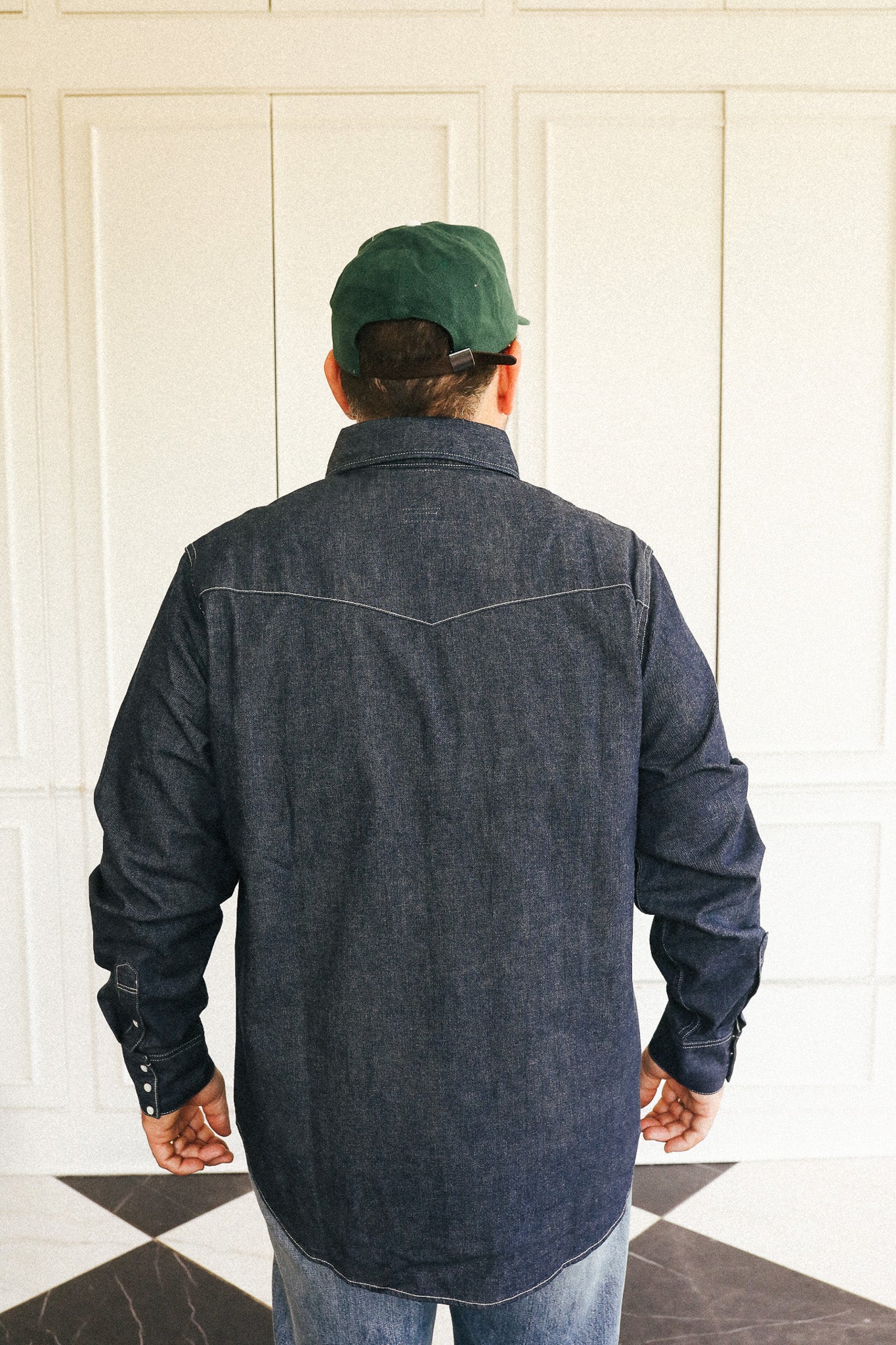 A man viewed from behind wearing a dark indigo denim long-sleeve shirt, blue jeans, and a green baseball cap, standing in front of a white paneled wall.