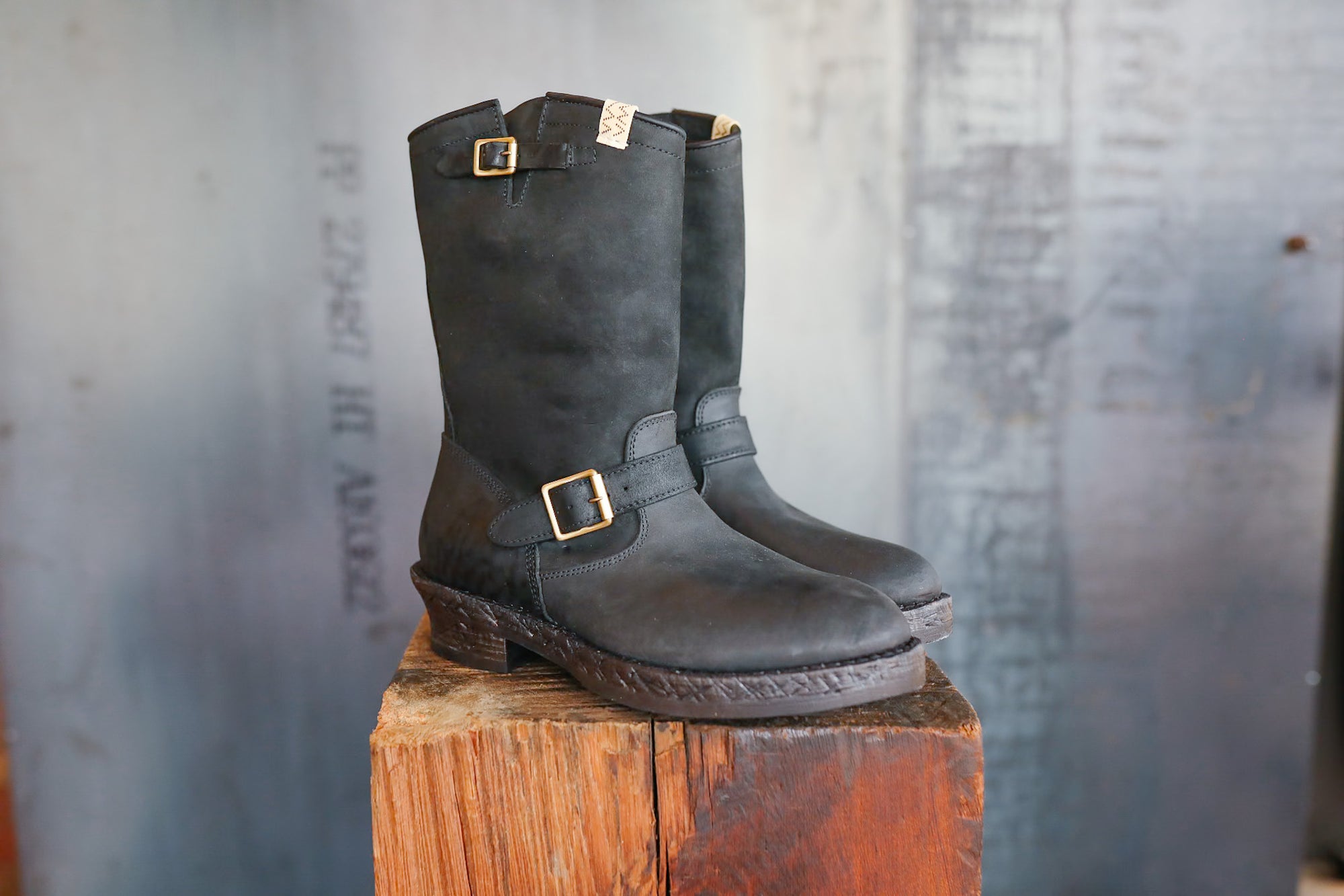 Pair of black leather boots with buckles on a wooden block against a textured wall.