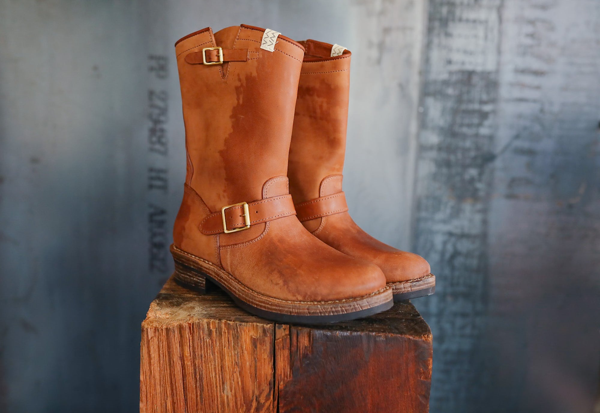 Pair of brown leather boots with buckles on a wooden block against a textured gray background