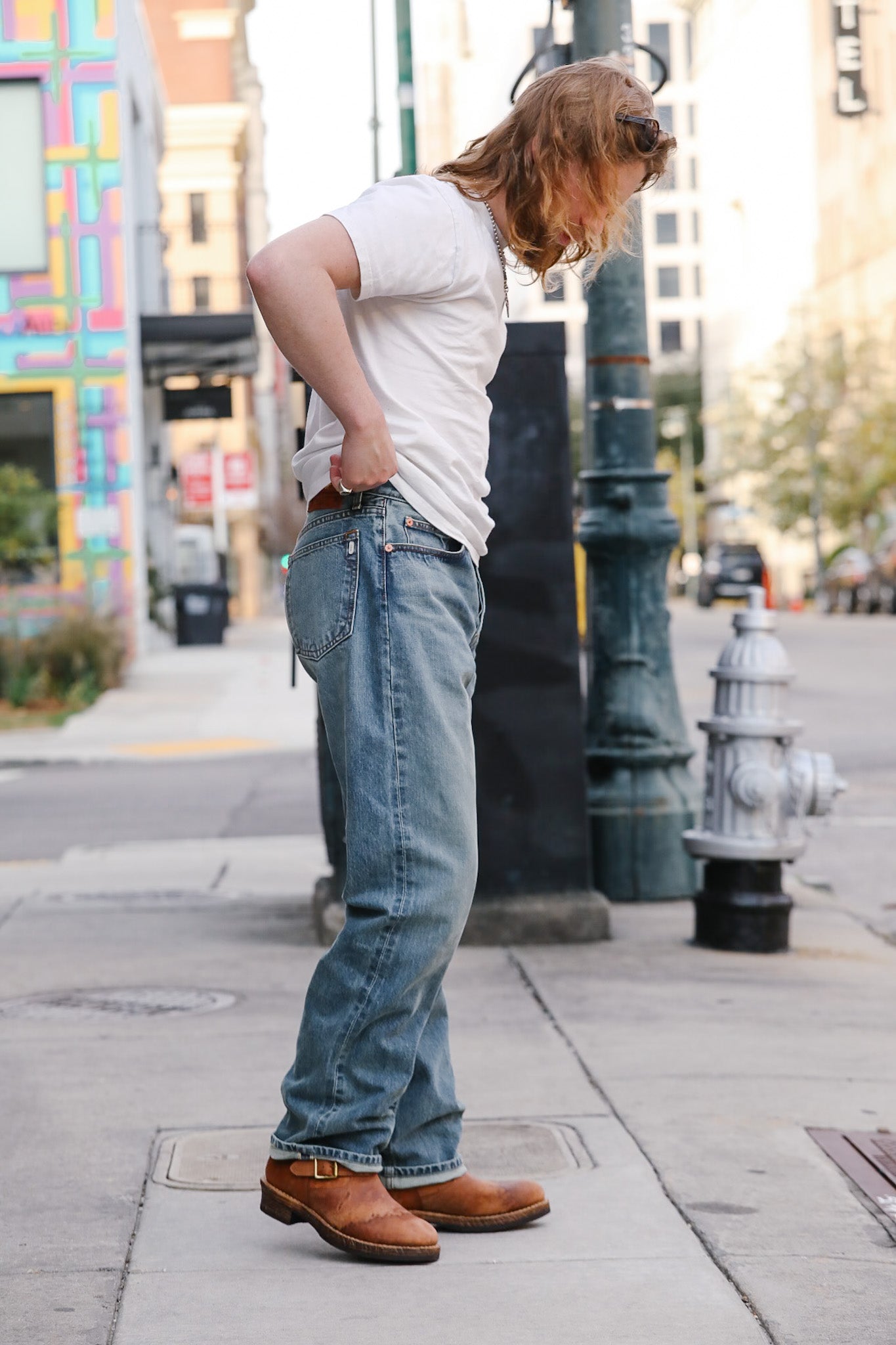 Person wearing a white sleeveless shirt, blue jeans, and brown shoes on a city street.