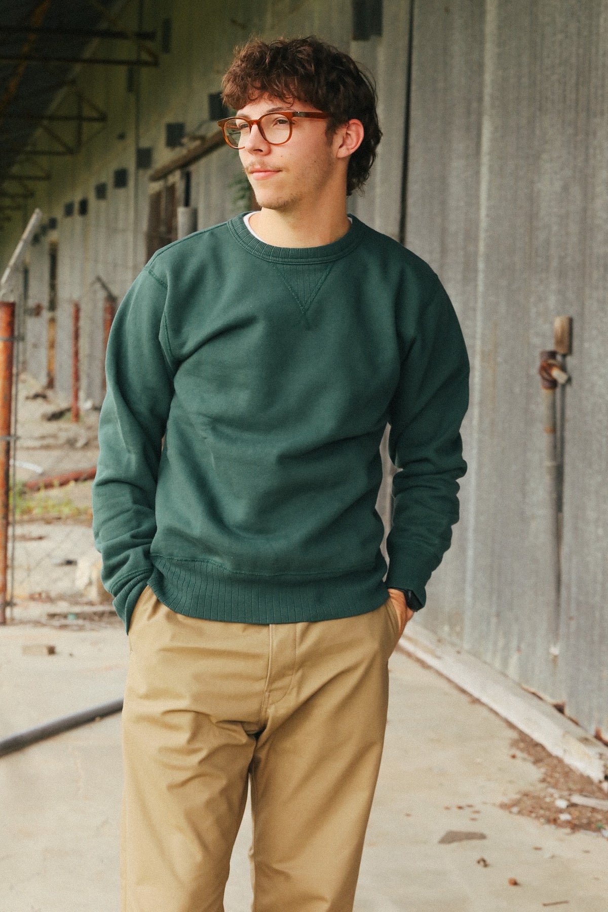 A man wearing a dark green crewneck sweatshirt and tan pants standing in an industrial corridor with a corrugated metal wall.