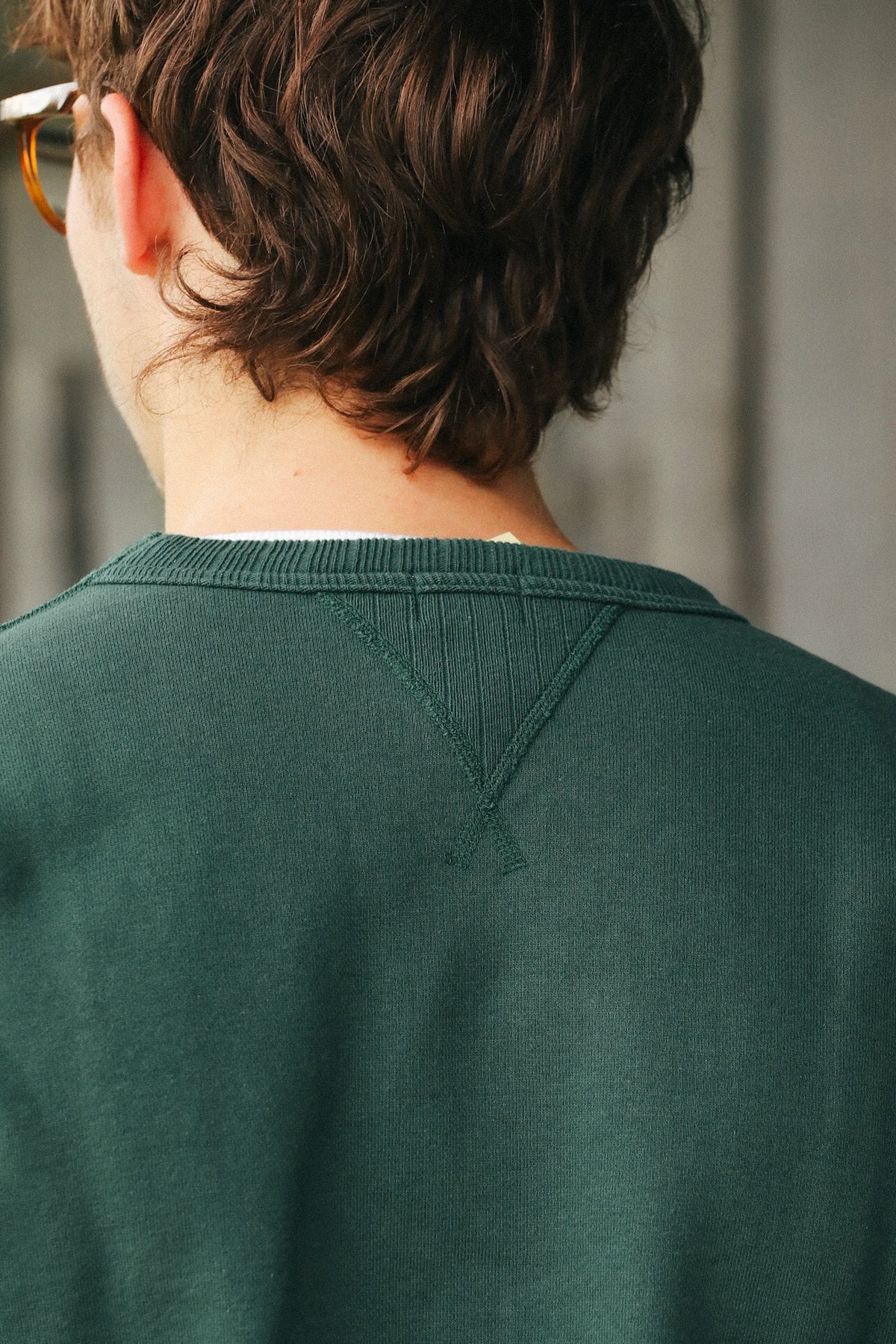 Close-up back view of a person wearing a dark green knit crewneck sweater, showing the V-neck detail and ribbed collar.