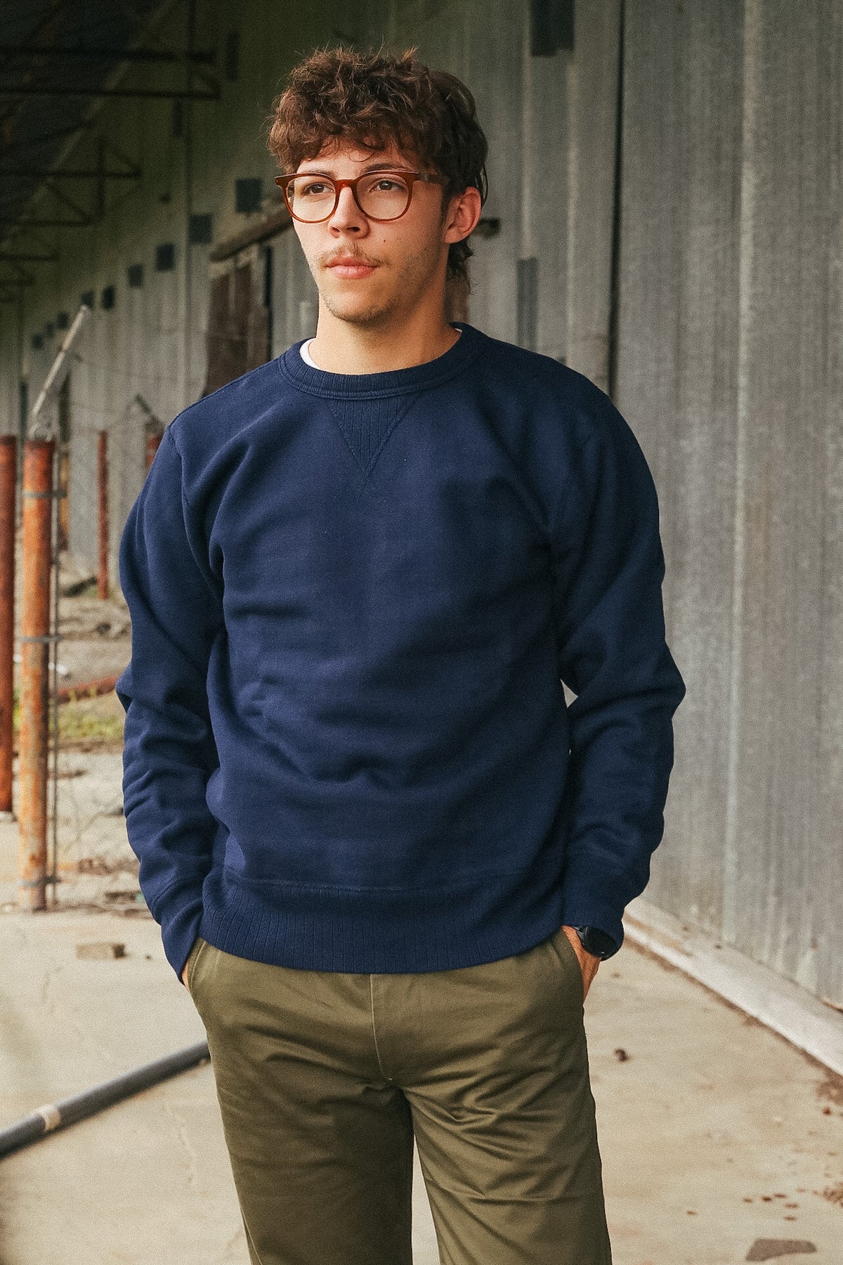 A man wearing a navy blue crewneck sweatshirt and olive green pants, standing in a concrete industrial corridor.