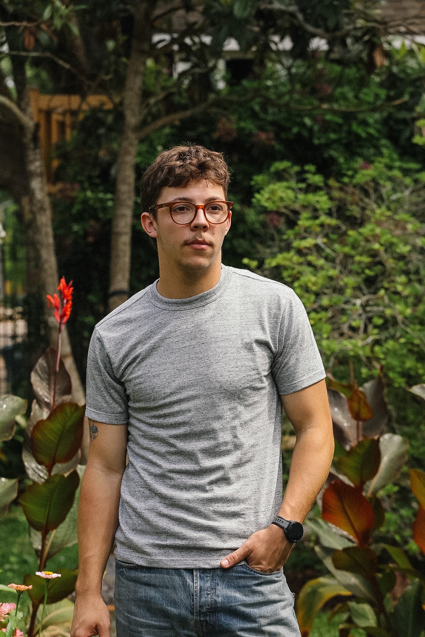 Man wearing a gray t-shirt and glasses standing in a garden with greenery.