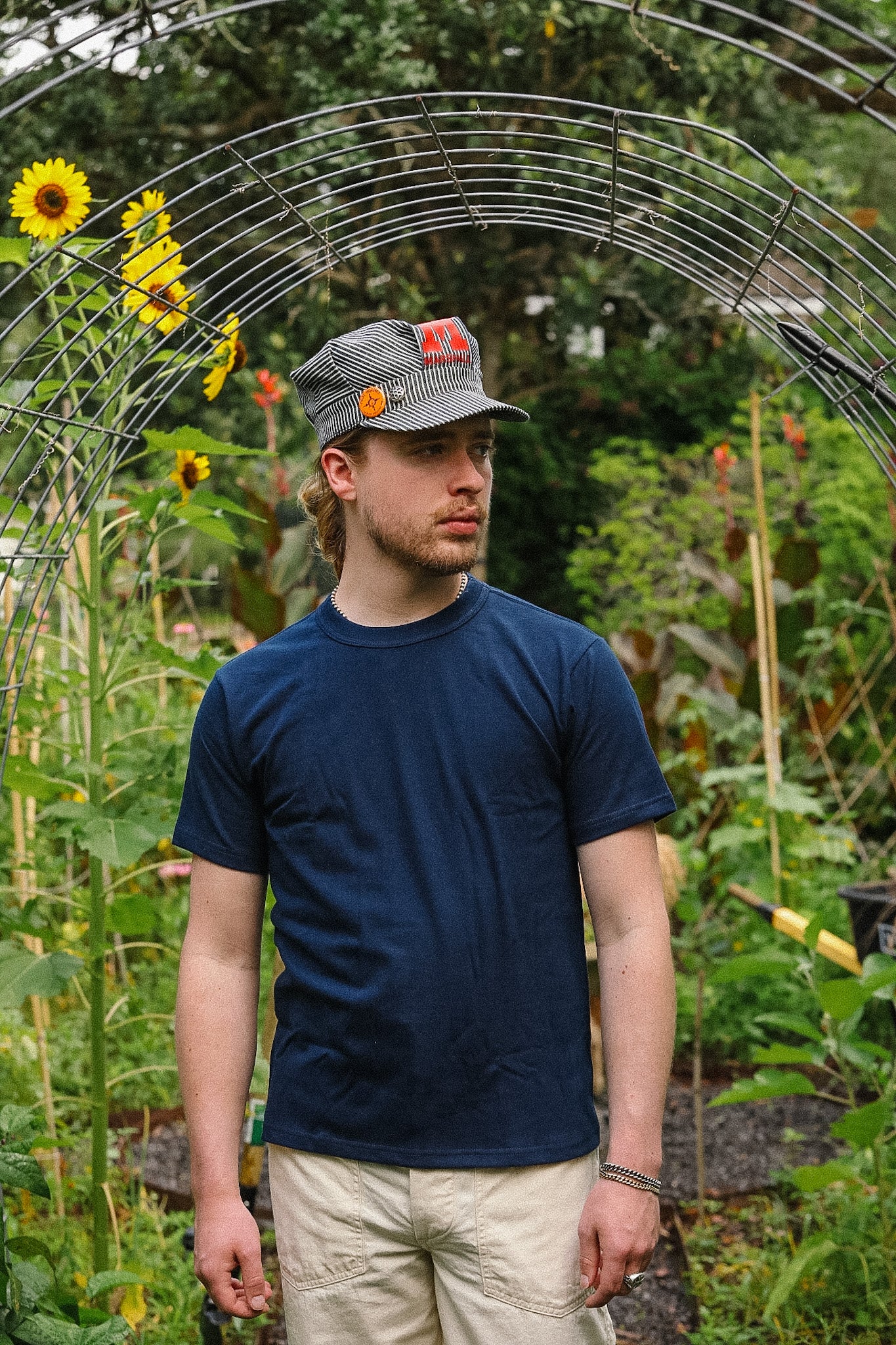 Man wearing a cap and blue shirt standing in a garden with sunflowers and greenery.