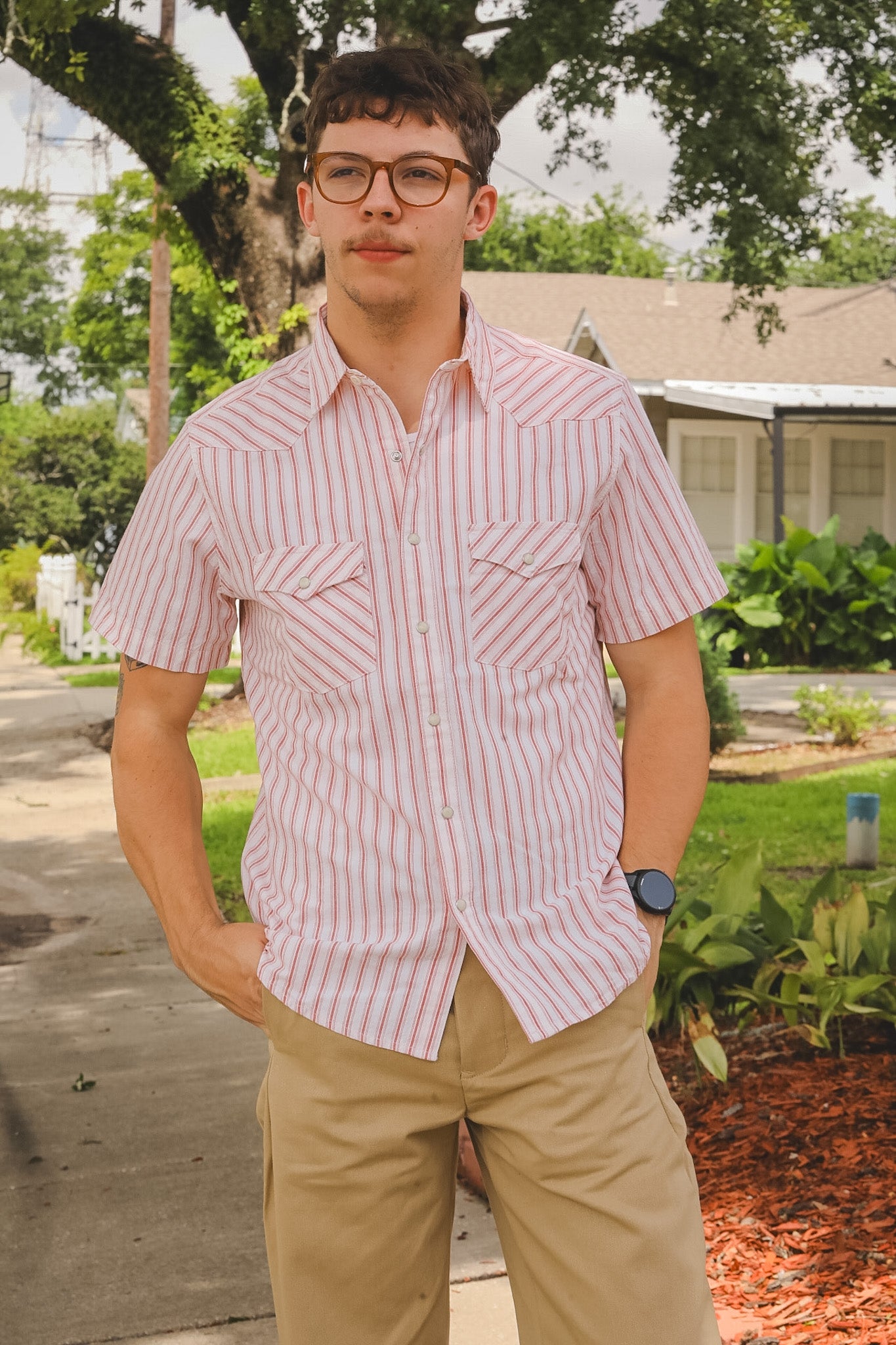 Man wearing a red and white striped shirt and beige pants standing outdoors.