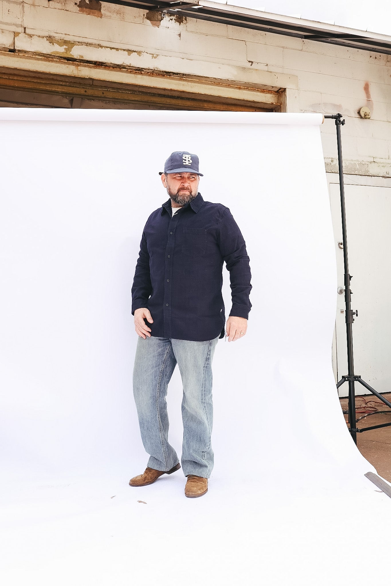 Man wearing a navy jacket, light gray jeans, and brown shoes standing against a white backdrop.
