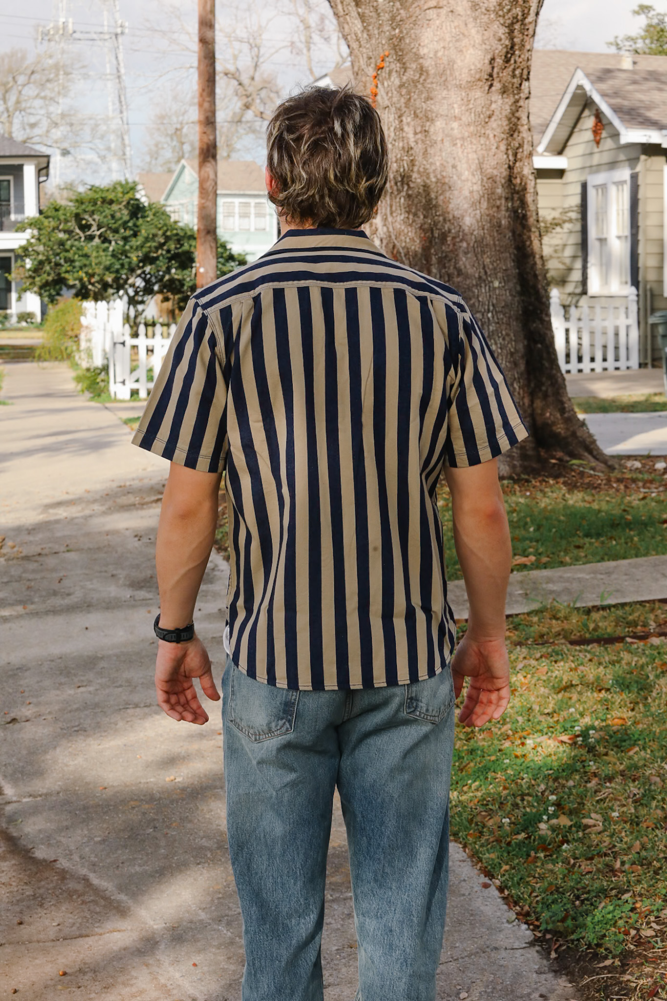 Man wearing a striped shirt and jeans walking on a sidewalk with houses and trees in the background.