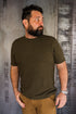 Bearded man wearing olive green short-sleeve shirt and tan pants against industrial backdrop