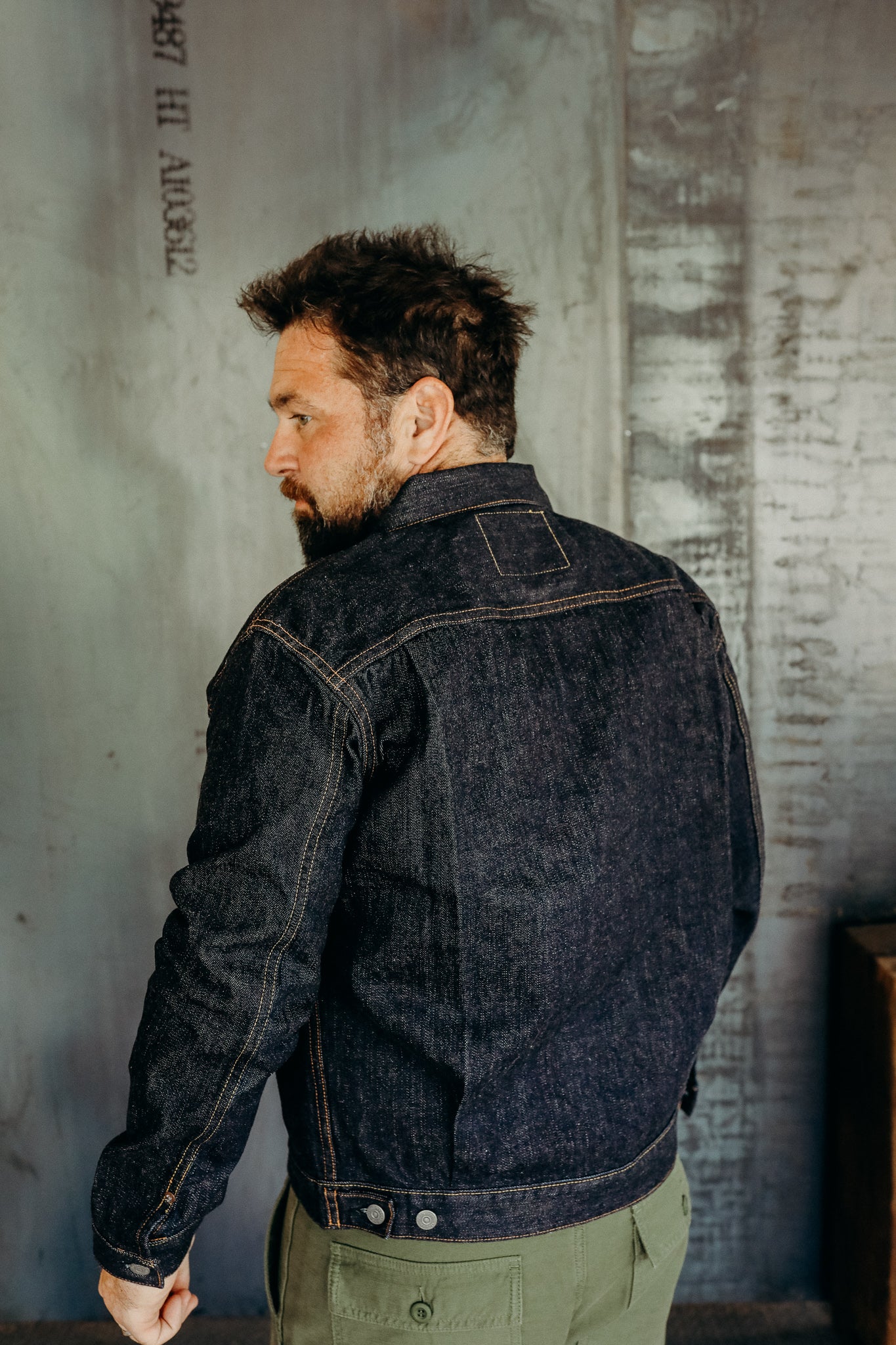 Man wearing dark denim jacket with contrast stitching standing indoors against textured wall
