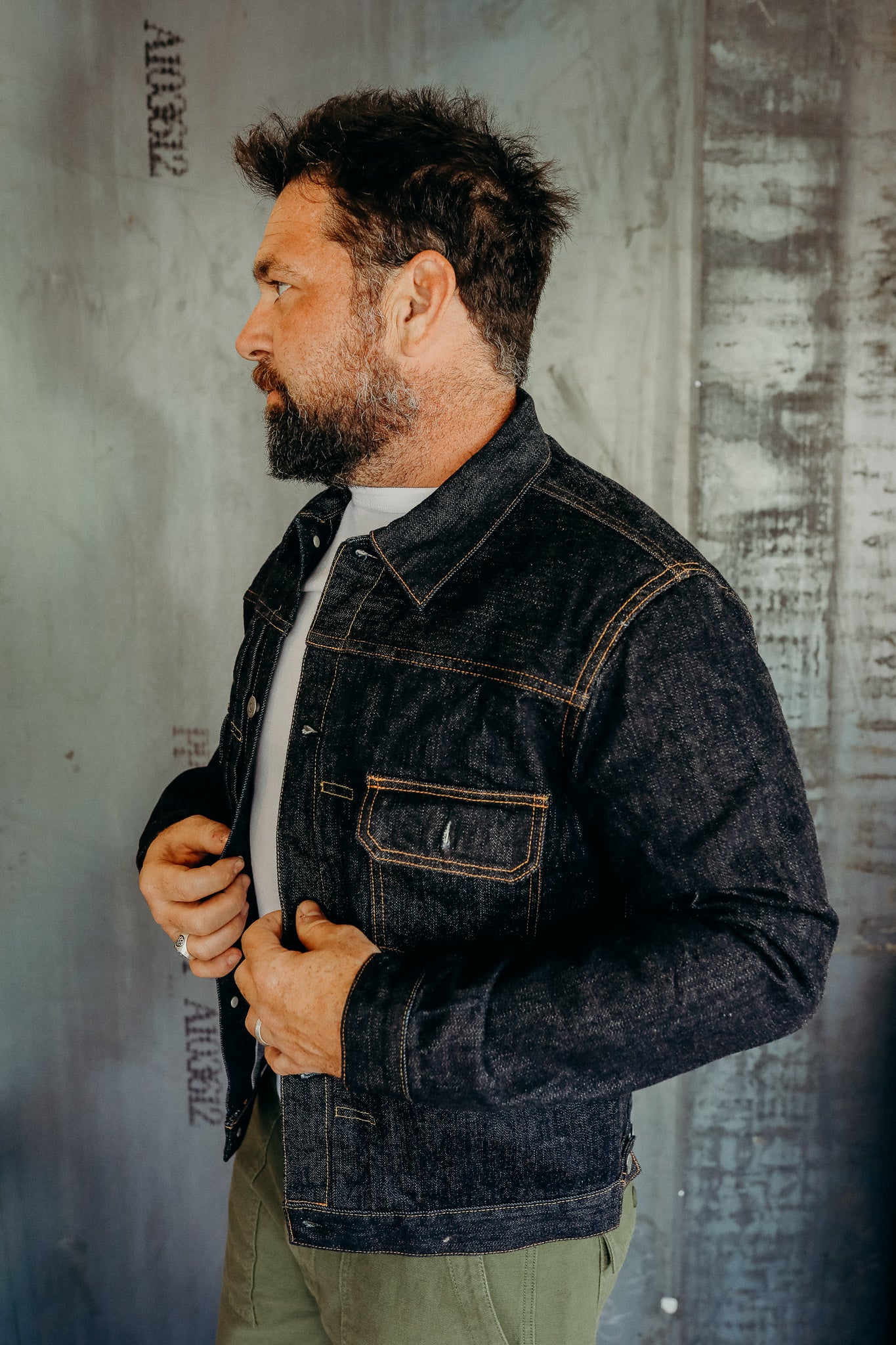 Man with beard wearing dark denim jacket over white shirt against industrial wall