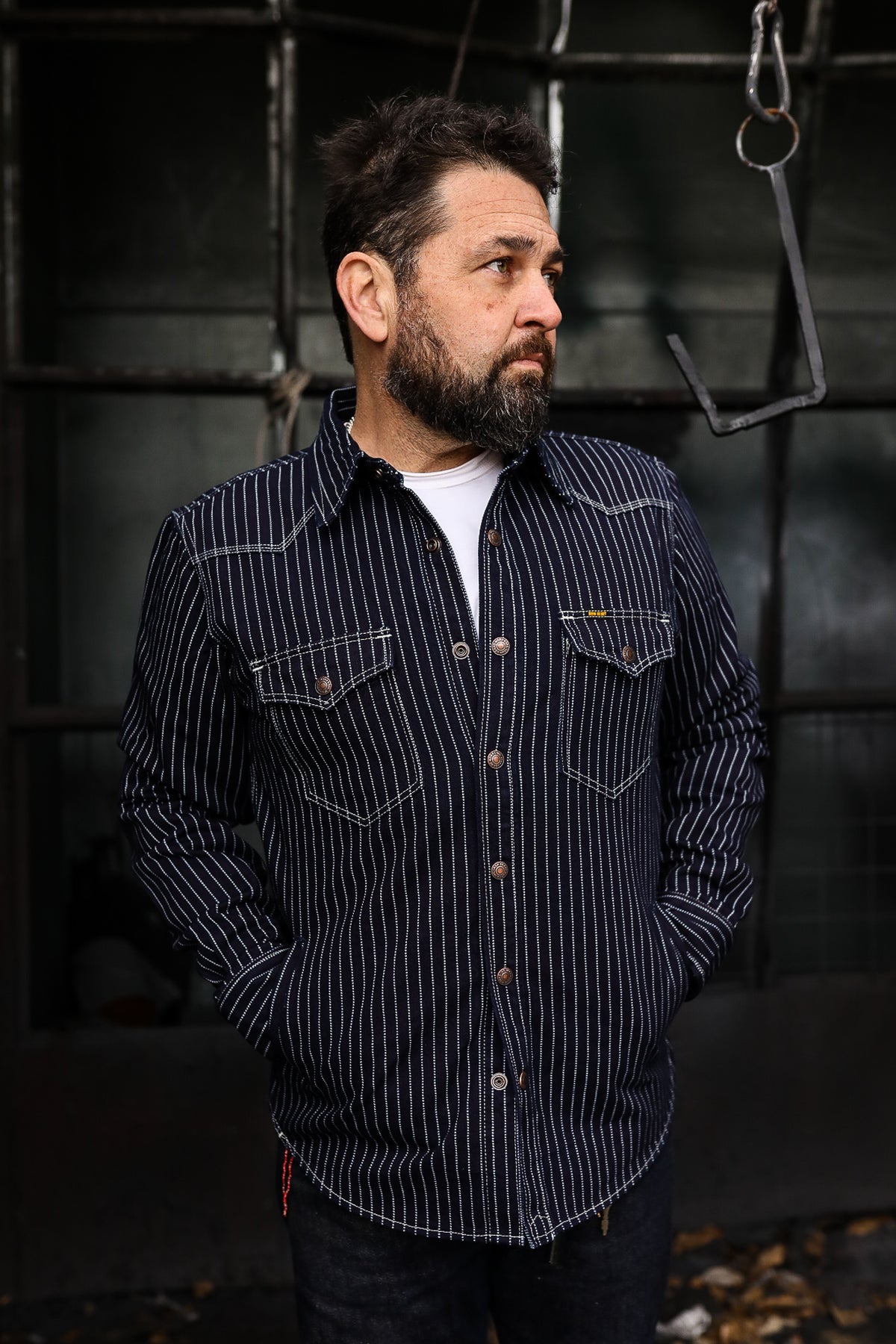 A person wearing a long-sleeved, striped, western-style CPO shirt with copper snap fastenings, standing against a industrial backdrop.
